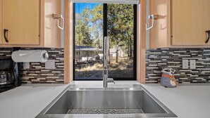 Une cuisine dotée d’un évier en acier inoxydable, de meubles en bois et offrant une vue sur un jardin avec des arbres et un barbecue.