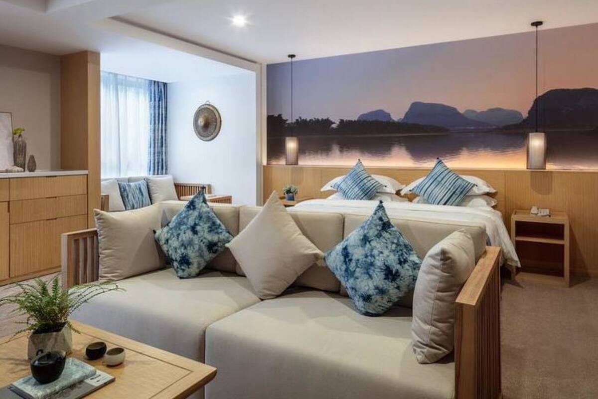 Une chambre d’hôtel moderne avec un grand lit, un canapé et une vue sur un lac au coucher du soleil.