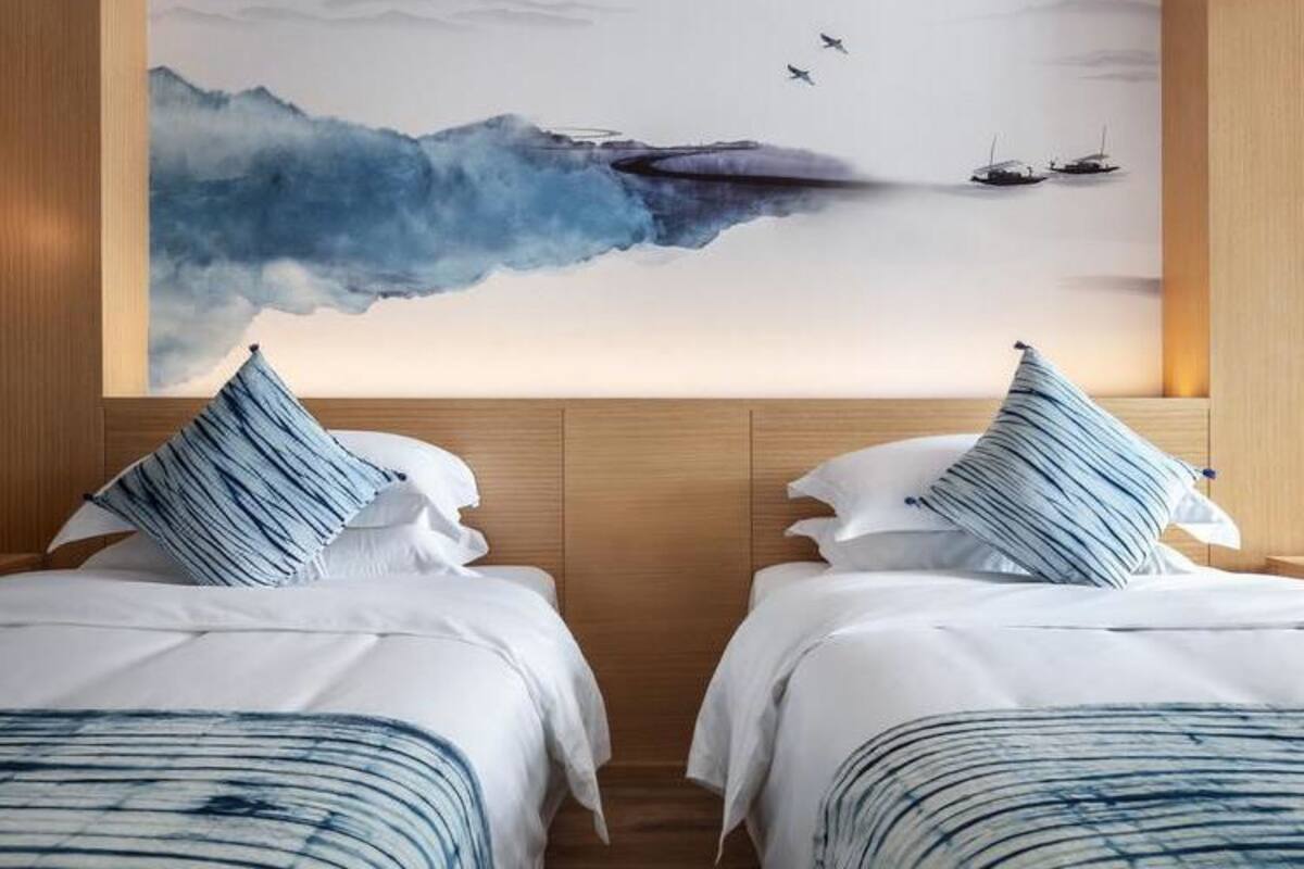Une chambre d’hôtel avec deux lits, chacun agrémenté d’un oreiller à rayures bleues, et une grande peinture murale représentant une mer calme et des bateaux.