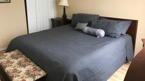 Ein ordentlich gemachtes Bett mit dunkelgrauer Bettdecke und Kissen, ein Nachttisch mit Lampe, eine blumengemusterte Bank und eine Tür mit Spiegel.
