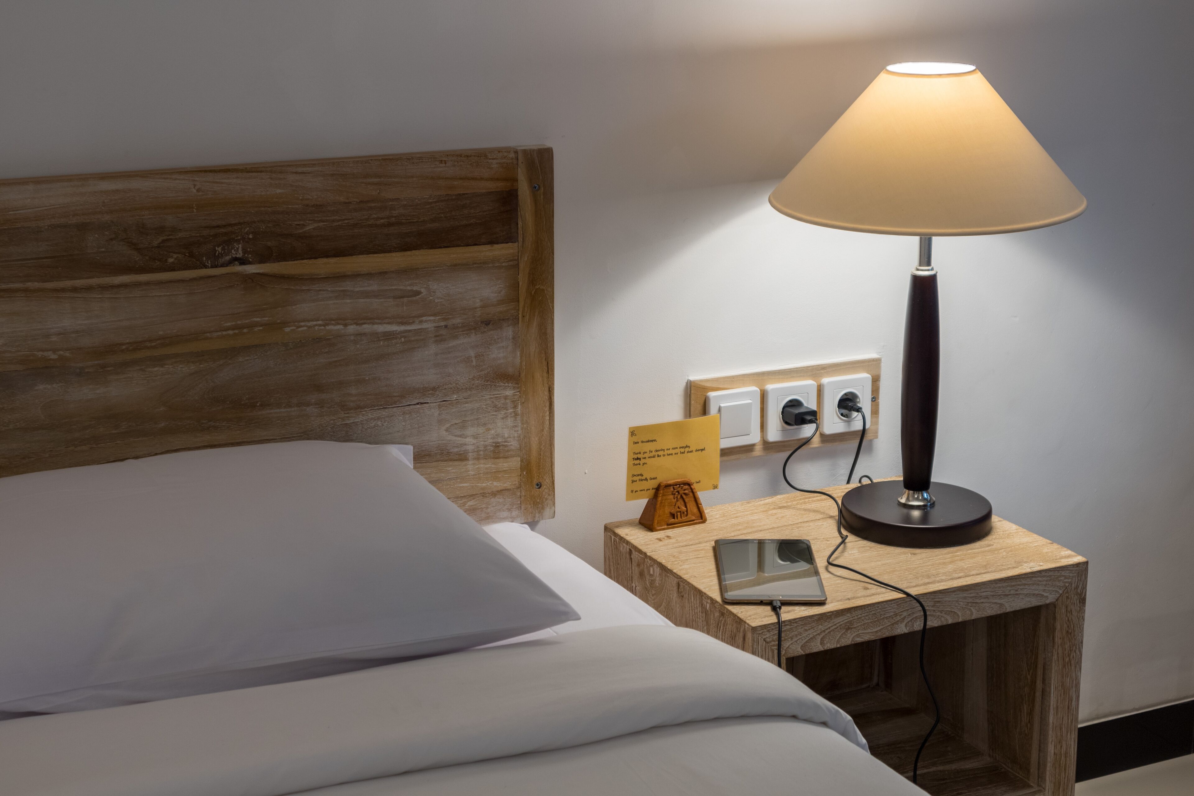 A neatly made bed with a wooden headboard, a bedside table with a lamp, a sign, and a smartphone.