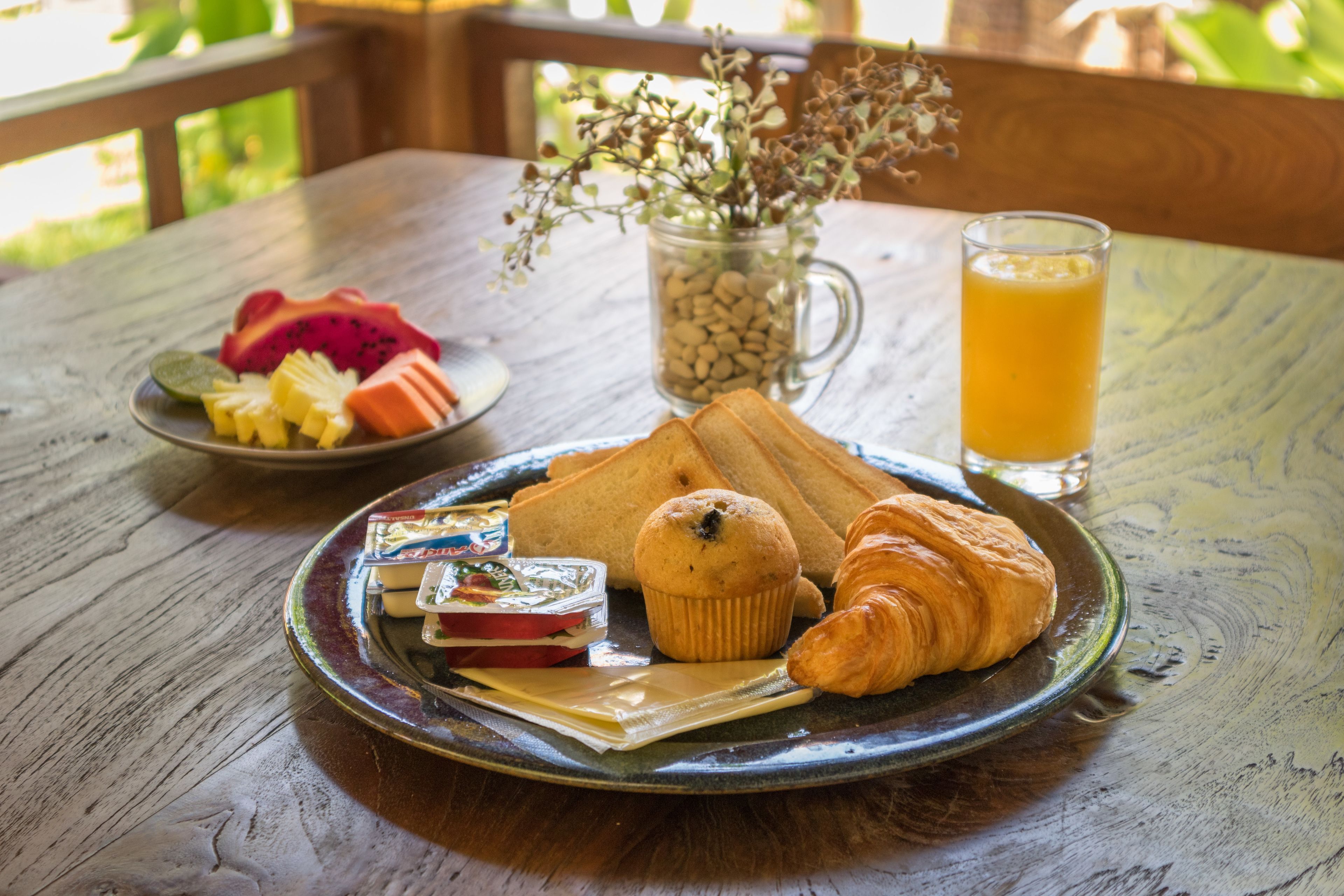 A breakfast setting with a plate of croissants, muffins, and fruit, a glass of orange juice, and a vase with dried flowers on a wooden table.
