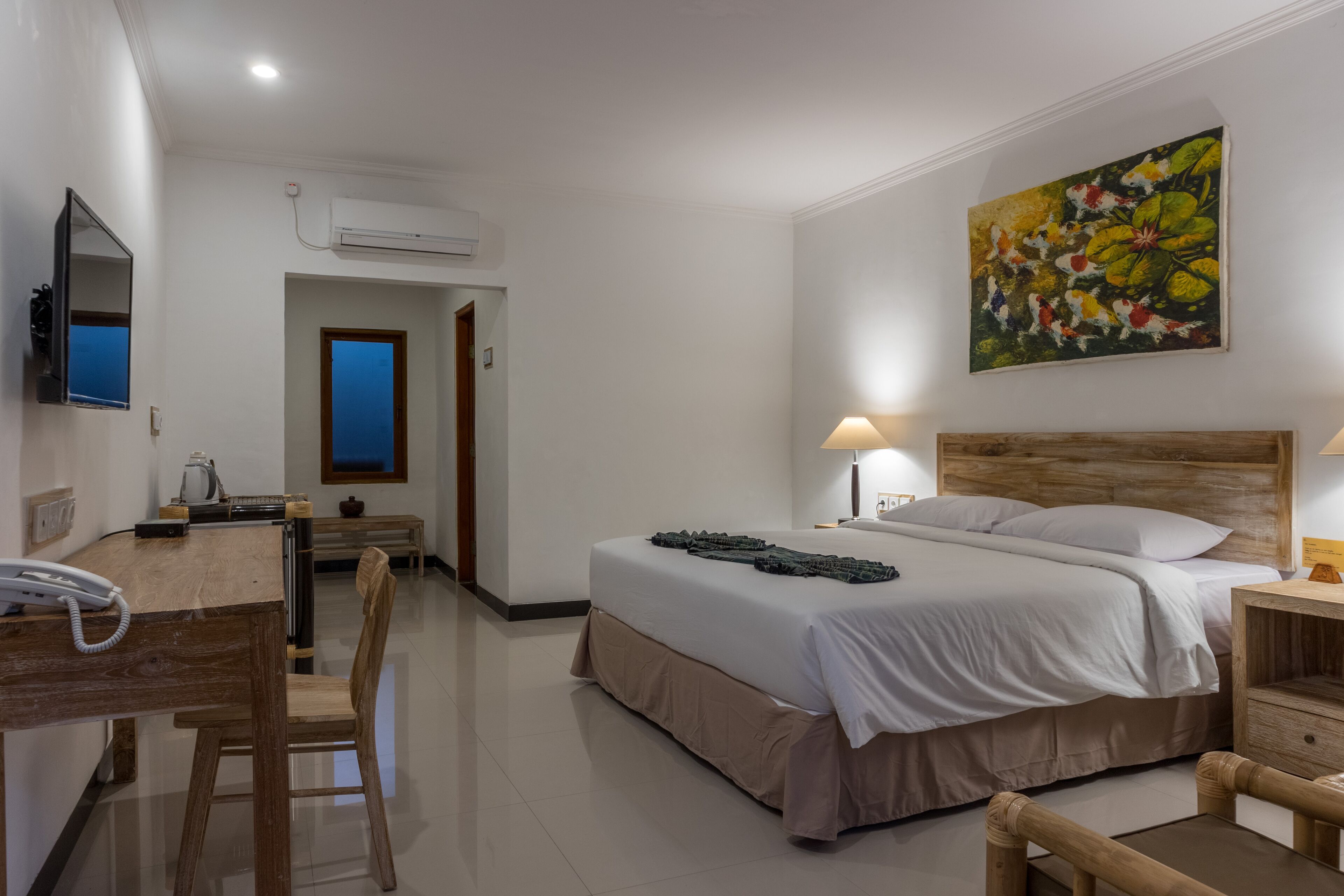 A hotel room with a bed, bedside table, lamp, desk with a telephone, and a painting on the wall.