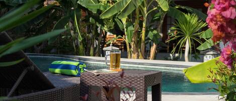 A poolside area with a wicker chair, a towel, a glass of beer, and lush tropical plants.