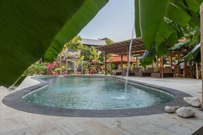 A swimming pool with a wooden deck, surrounded by lush greenery and a covered seating area. - Sanur Lodge (Denpasar)