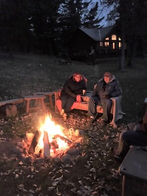 Een kampvuur met mensen eromheen zittend, een houten bankje en een huis op de achtergrond.