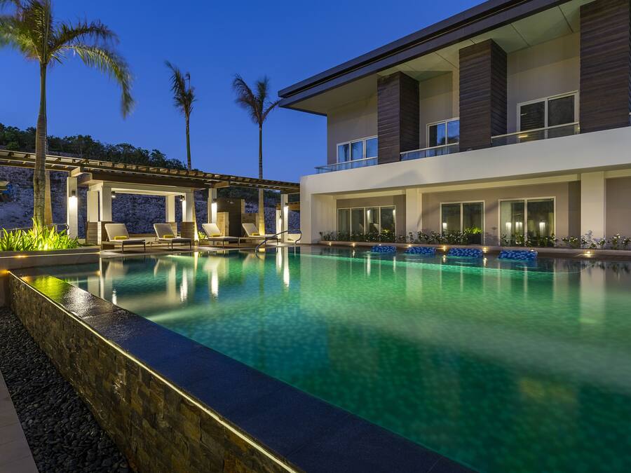 Um hotel moderno com piscina, espreguiçadeiras e uma área coberta com palmeiras.
