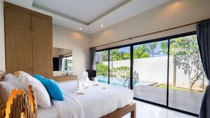 A modern bedroom with a large bed, a wooden wardrobe, and a view of a swimming pool and greenery outside. - Layantara Villas (Thep Krasattri)