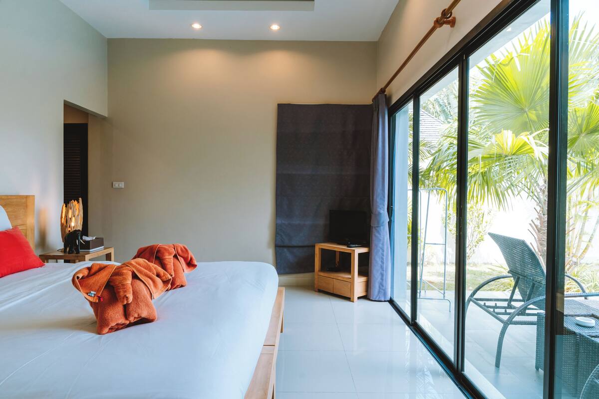 A modern hotel room with a large bed, a small table, and a view of palm trees through large windows.