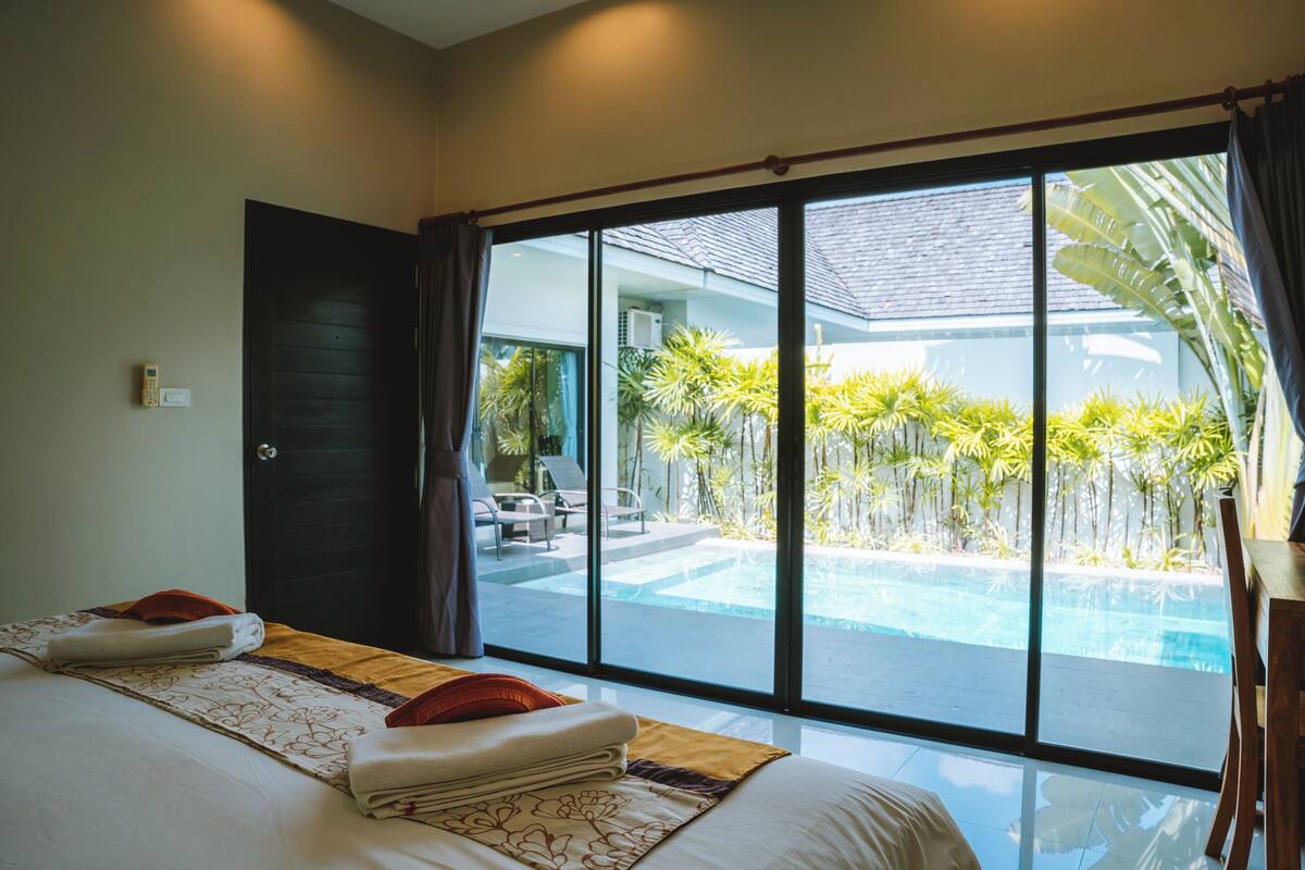 A hotel room with a large sliding glass door leading to a pool area, a bed with neatly folded towels, and a view of the pool and surrounding greenery.