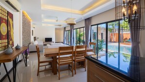 A modern dining area with a wooden table and chairs, a TV in the background, and a view of a swimming pool. - Layantara Villas (Thep Krasattri)