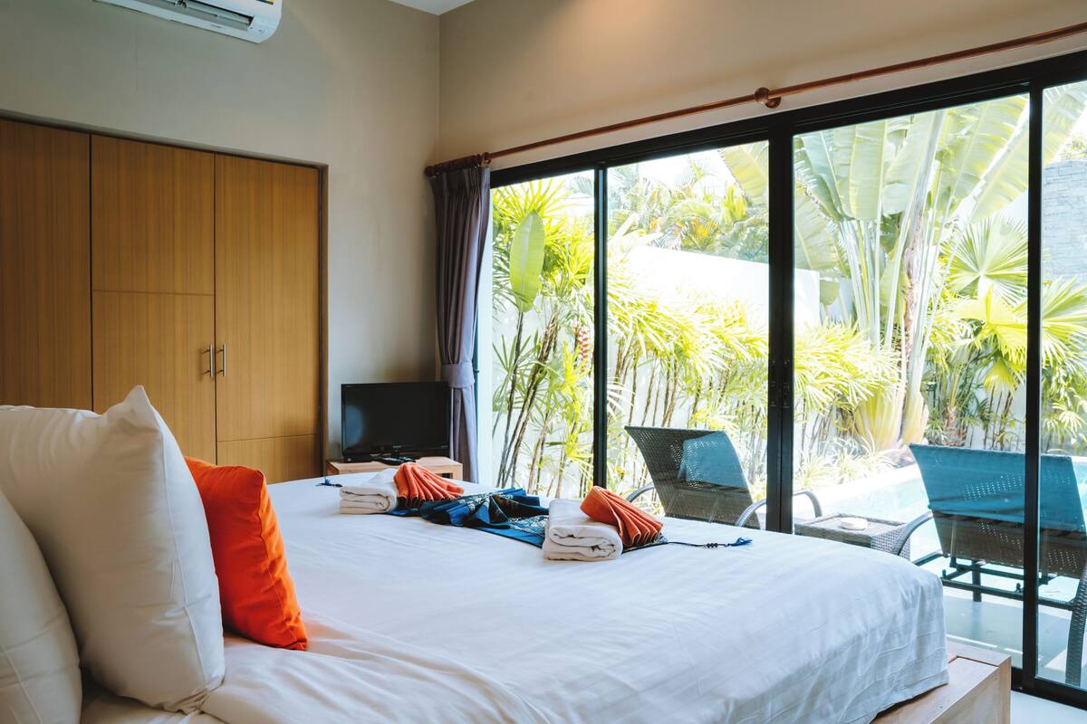 A hotel room with a large bed, a TV, and a view of a pool and tropical plants.