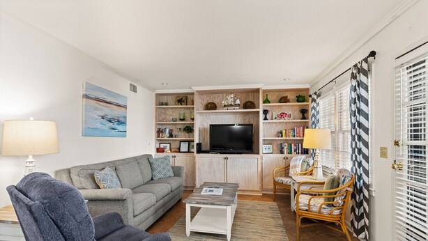 A living room with a grey sofa, a blue armchair, a coffee table, and a TV. There are shelves with books and decorative items, and a window with blinds. - Comfortable 2 bedroom Beach Home located on quiet street (Saint Simons Island)