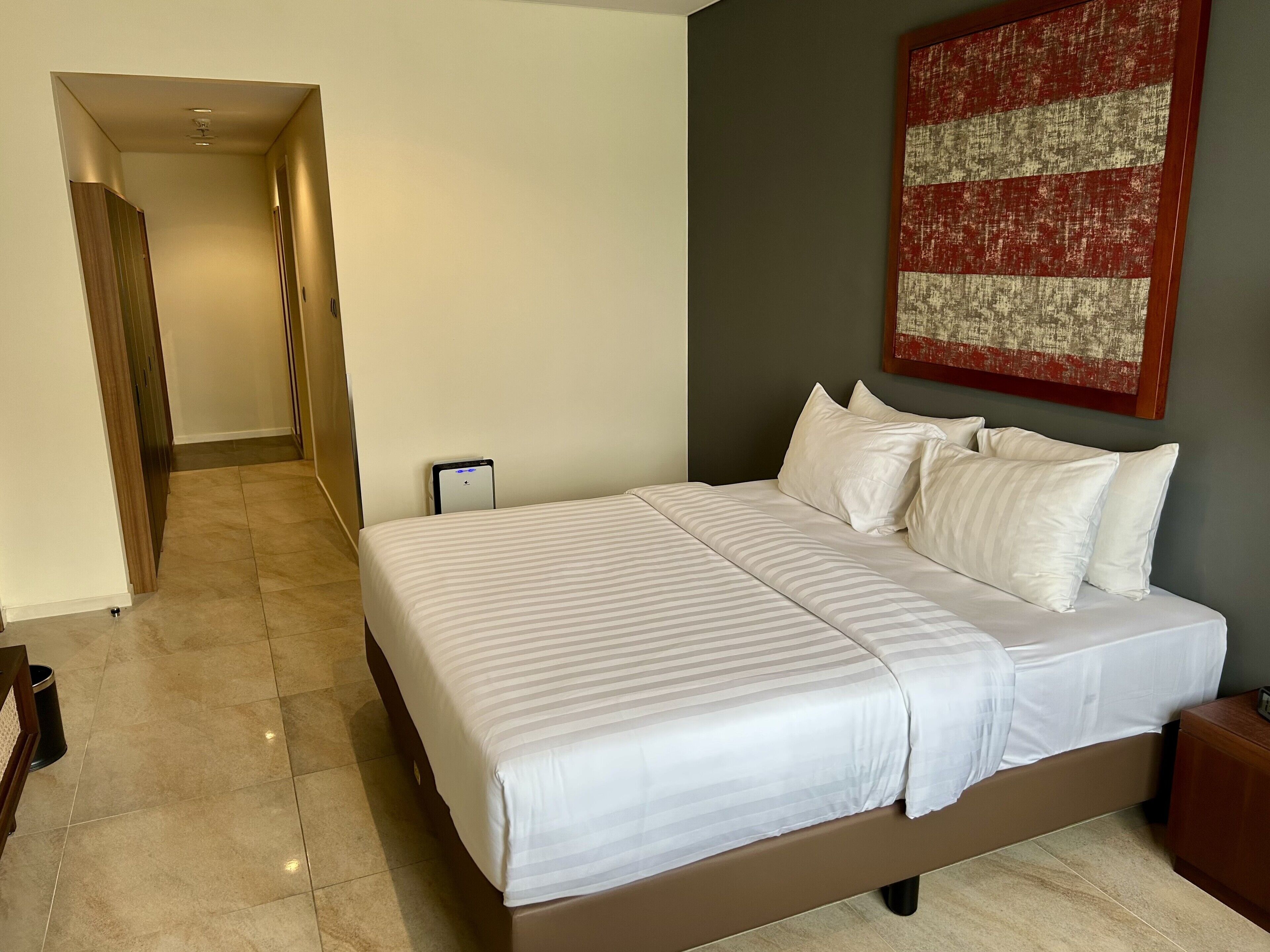 Ein Hotelzimmer mit einem großen Bett, einem Nachttisch, einem Wandbild und einem Flur, der in einen anderen Raum führt.