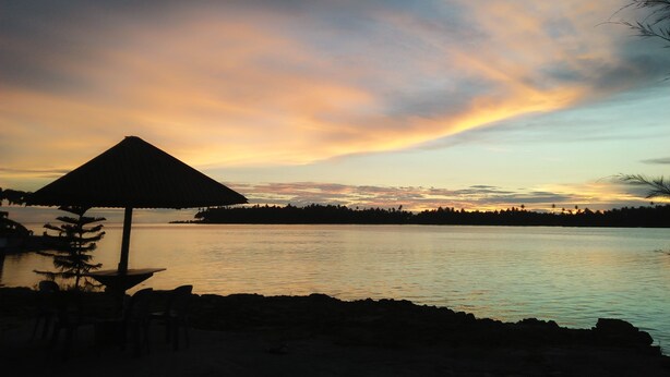 A beach view with a thatched umbrella and chairs, a calm lake, and a sunset sky. - Salty Dog Hostel (Nias)