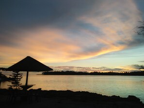 Une plage avec une vue sur un lac paisible, un parasol en chaume et des chaises, et un ciel au coucher du soleil.