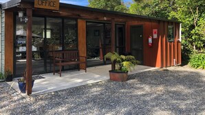 A small wooden building labeled "OFFICE" with a bench outside, a fire extinguisher, and a gravel driveway. - Kanuka Ridge Lodge and Backpackers (Kaiteriteri)