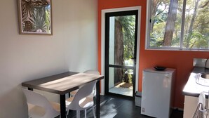 A compact kitchen with a dining area, a refrigerator, and a window with a view of trees. - Kanuka Ridge Lodge and Backpackers (Kaiteriteri)