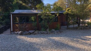 A small, rustic cabin with a metal roof, surrounded by gravel and vegetation. - Kanuka Ridge Lodge and Backpackers (Kaiteriteri)
