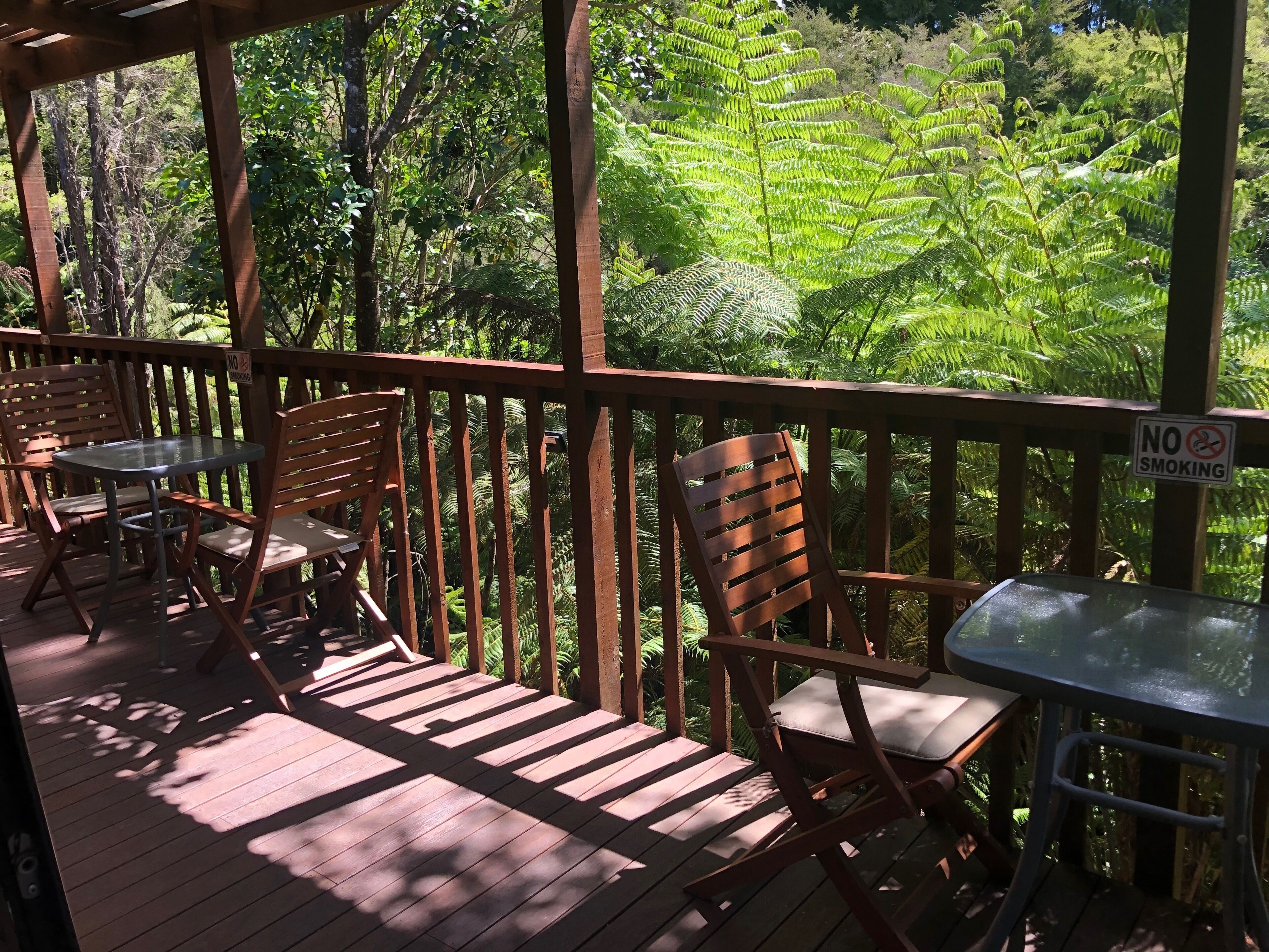 A wooden deck with outdoor seating, including chairs and a table, surrounded by lush greenery and ferns.