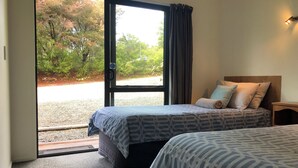 A hotel room with two beds, a view of trees and a gravel path through the door, and a nightstand with a lamp. - Kanuka Ridge Lodge and Backpackers (Kaiteriteri)