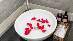 Egy fehér mosdókagyló, rózsalapok, két üveg sampon és egy kis kosár, tele piperecikkekkel.