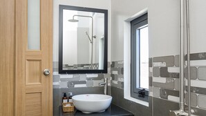 Egy modern fürőszoba, melyben fa ajtó, fehér mosdókagyló, tükör és zuhanyzó található.