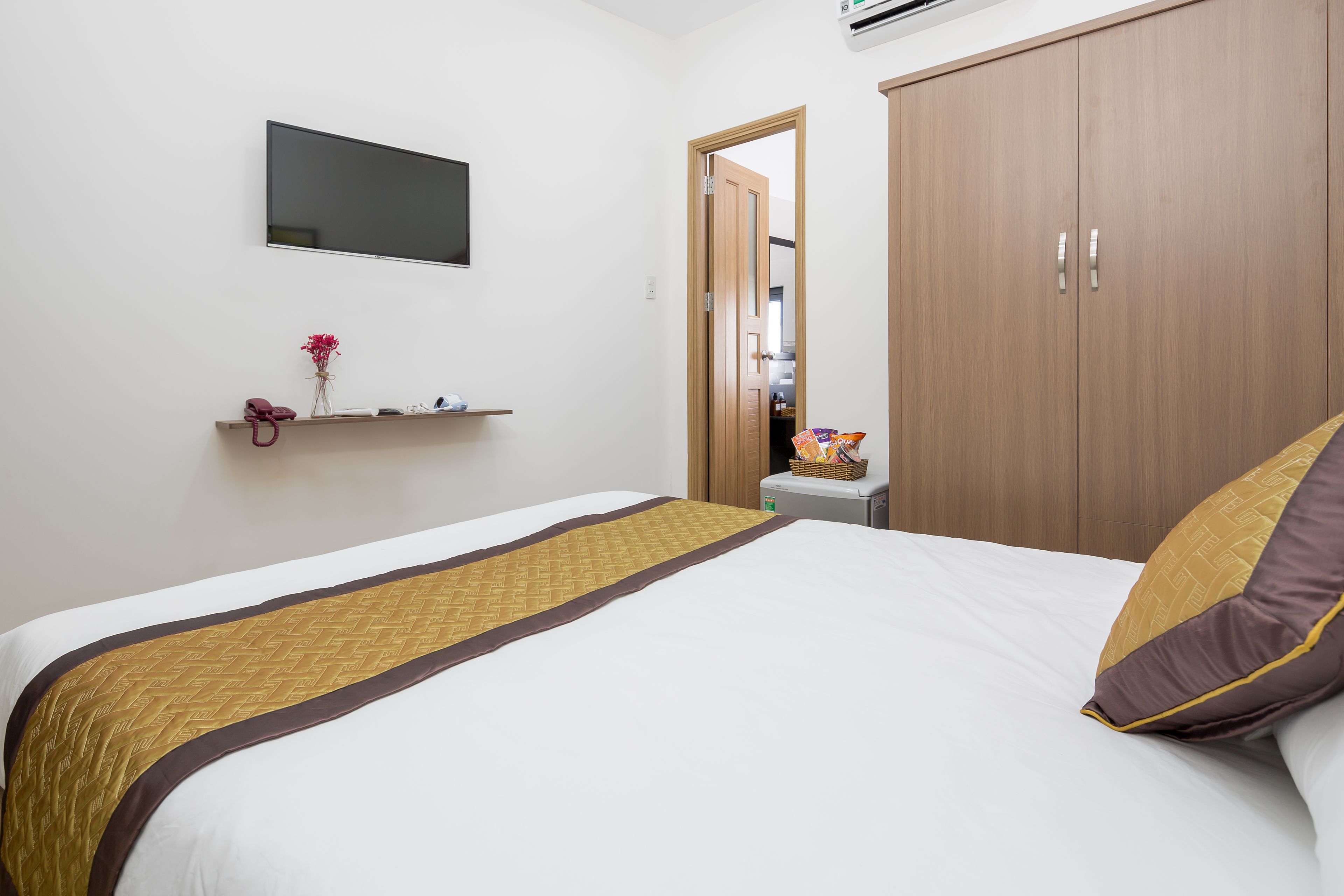 A hotel room with a bed, a TV mounted on the wall, a wooden wardrobe, and a small table with a basket.