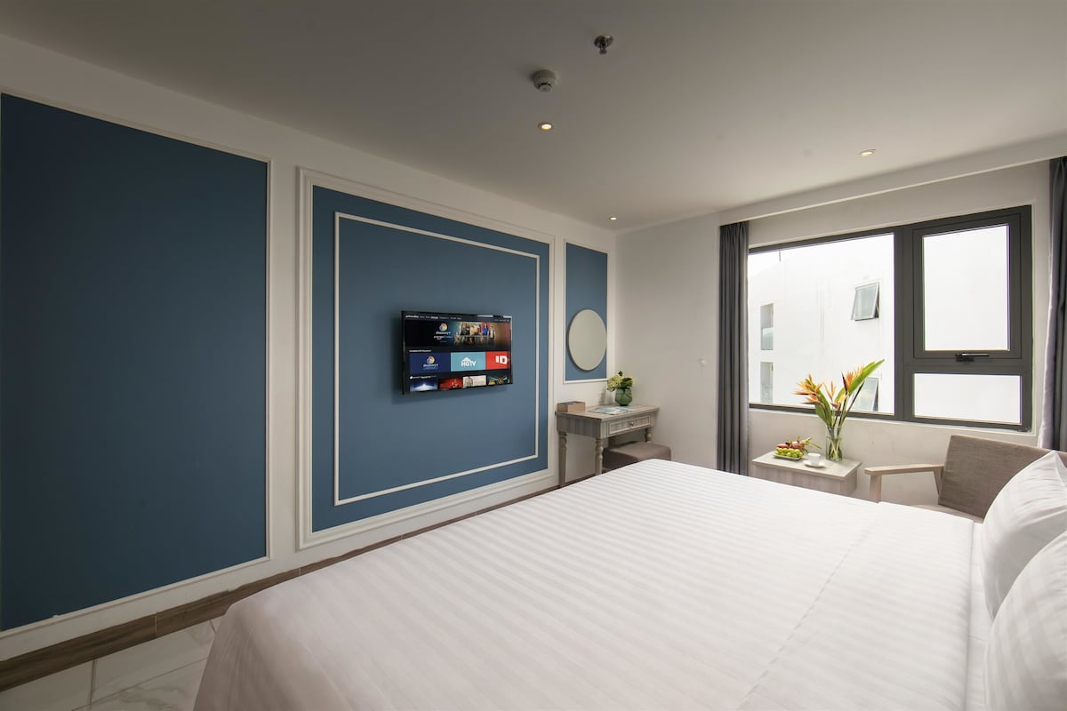 Habitació d'hotel amb un llit gran, una televisió penjada a la paret, una taula petita amb un vas de flors i una finestra amb vista a edificis.
