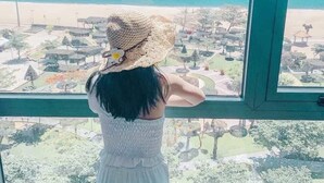 A person in a white dress and straw hat looking out from a window at a coastal view with a beach and a park. - Ruby Luxury Hotel (Nha Trang)