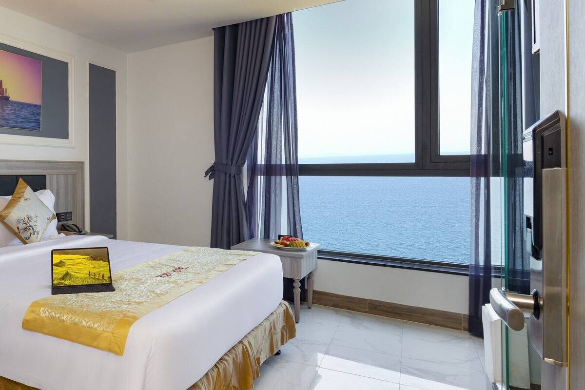 A hotel room with a bed, a laptop, a small table with fruit, and a view of the sea through large windows.