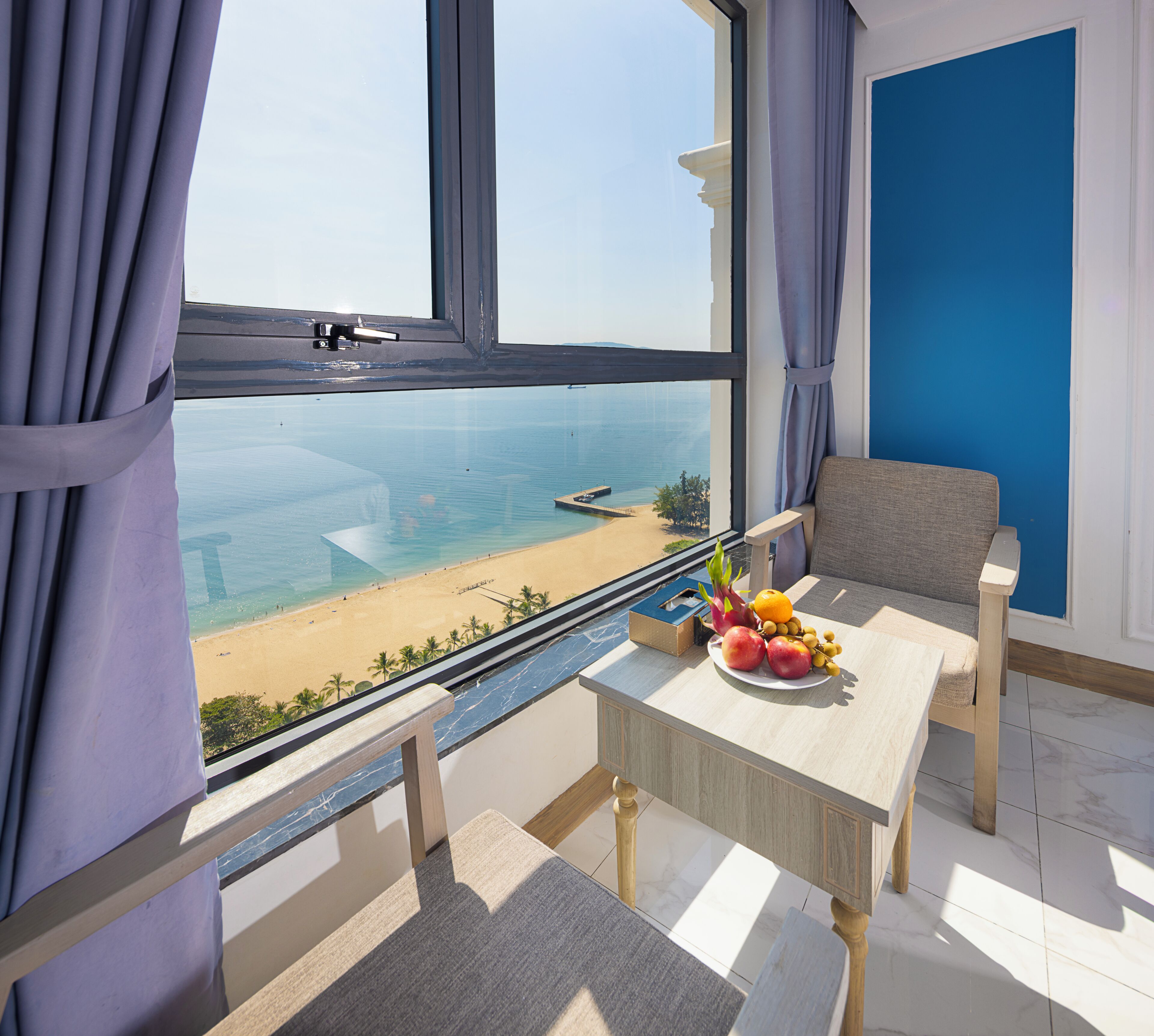 A room with a view of a beach, a table with fruit, and a chair.