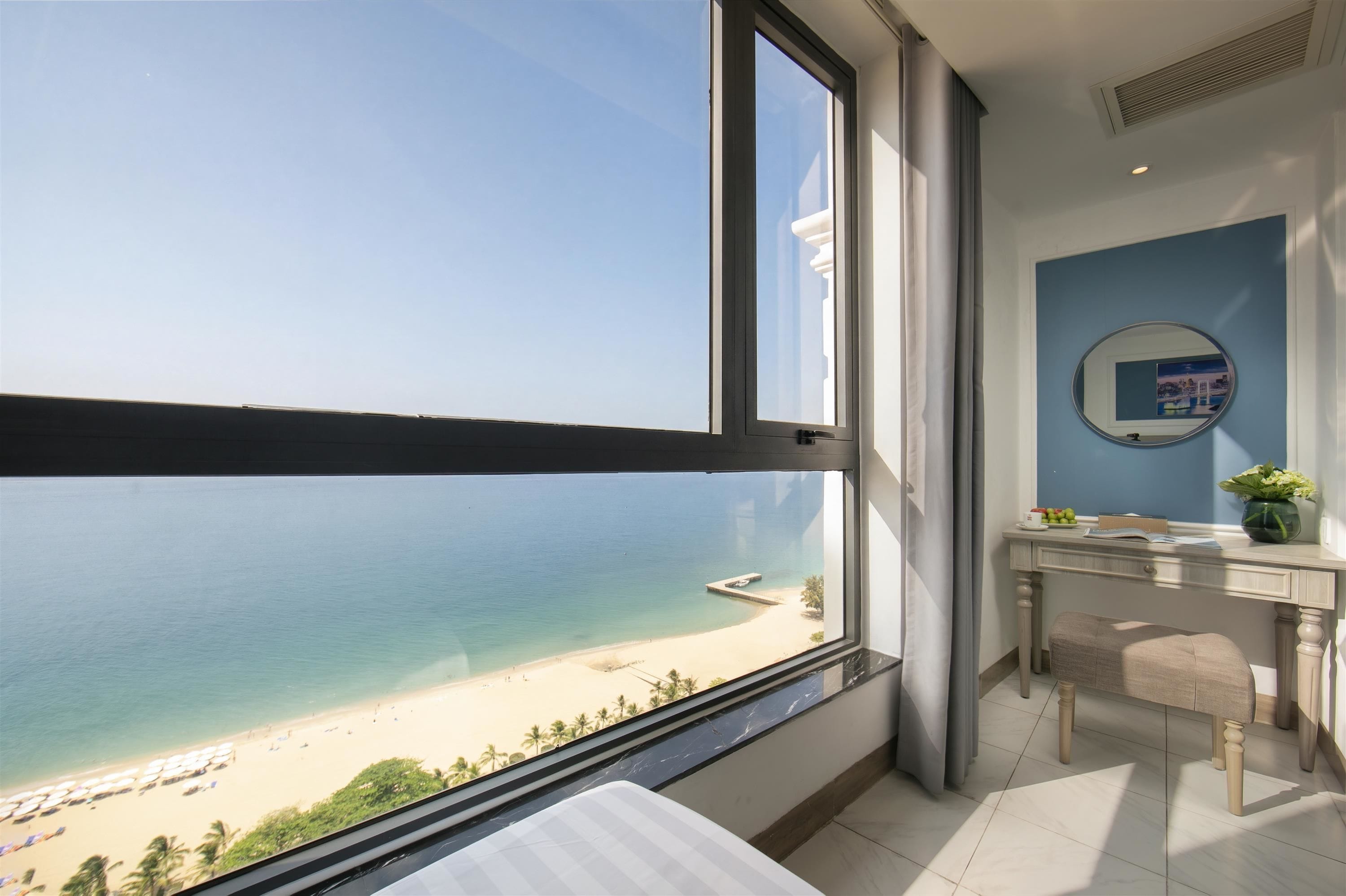 A room with a large window overlooking a beach, a bathroom with a mirror, and a desk with a vase of flowers.