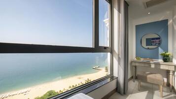 A room with a large window overlooking a beach, a bathroom with a mirror, and a desk with a vase of flowers.