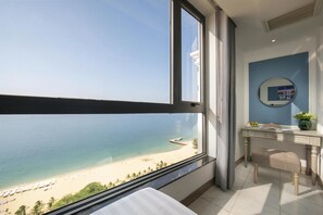 A room with a large window overlooking a beach, a bathroom with a mirror, and a desk with a vase of flowers. - Ruby Luxury Hotel (Nha Trang)