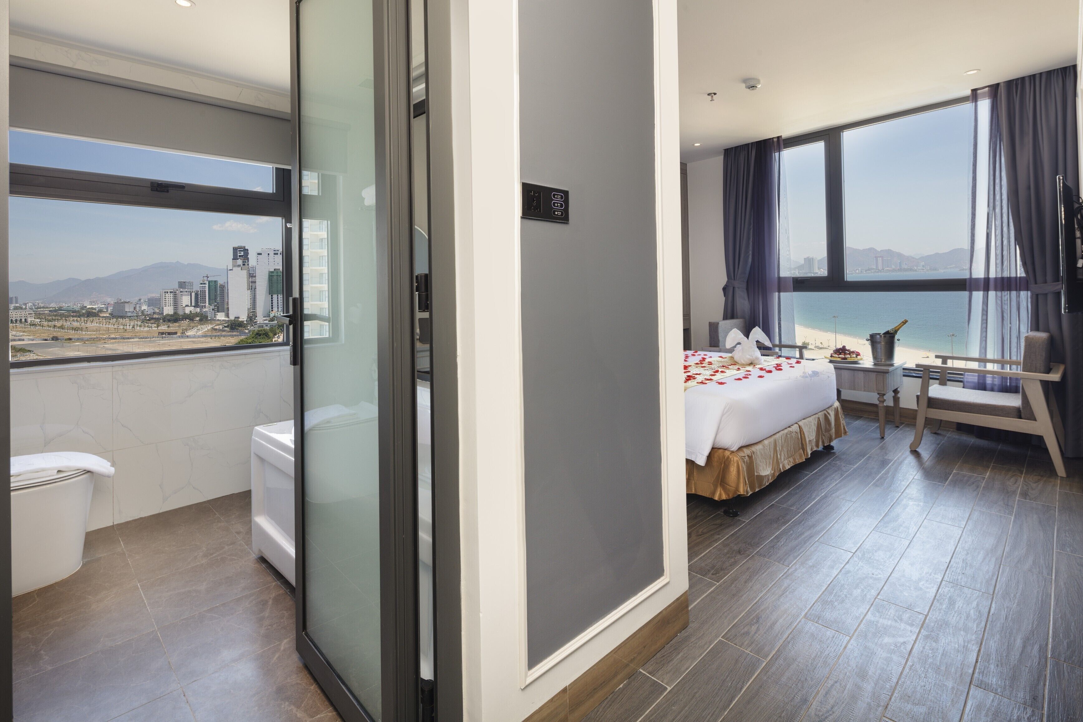 A hotel room with a balcony, a bed, a chair, and a view of the city and beach.