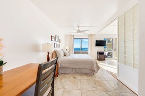 A hotel room with a bed, a desk, a chair, a ceiling fan, a TV, and a view of the ocean.