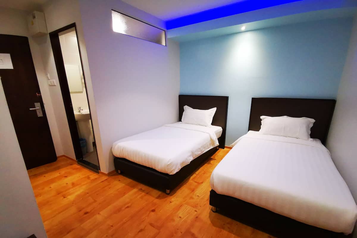 A hotel room with two single beds, a wooden floor, a small bathroom with a sink, and a door leading to another room.
