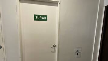 A door labeled "SURAU" with a doormat, a vent, and a bench with a bag on it.