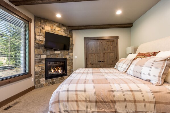 A bedroom with a stone fireplace, a bed with plaid bedding, a wooden door, and a TV mounted on the wall. - Sunstone Retreat with Access to Forest House (Bend)