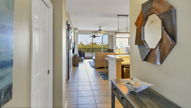 A hallway with a view to a balcony, a dining area, and a kitchen with a large mirror and a bowl of fruit.