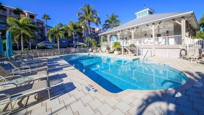A swimming pool with lounge chairs and umbrellas, surrounded by palm trees and a building with a covered area.