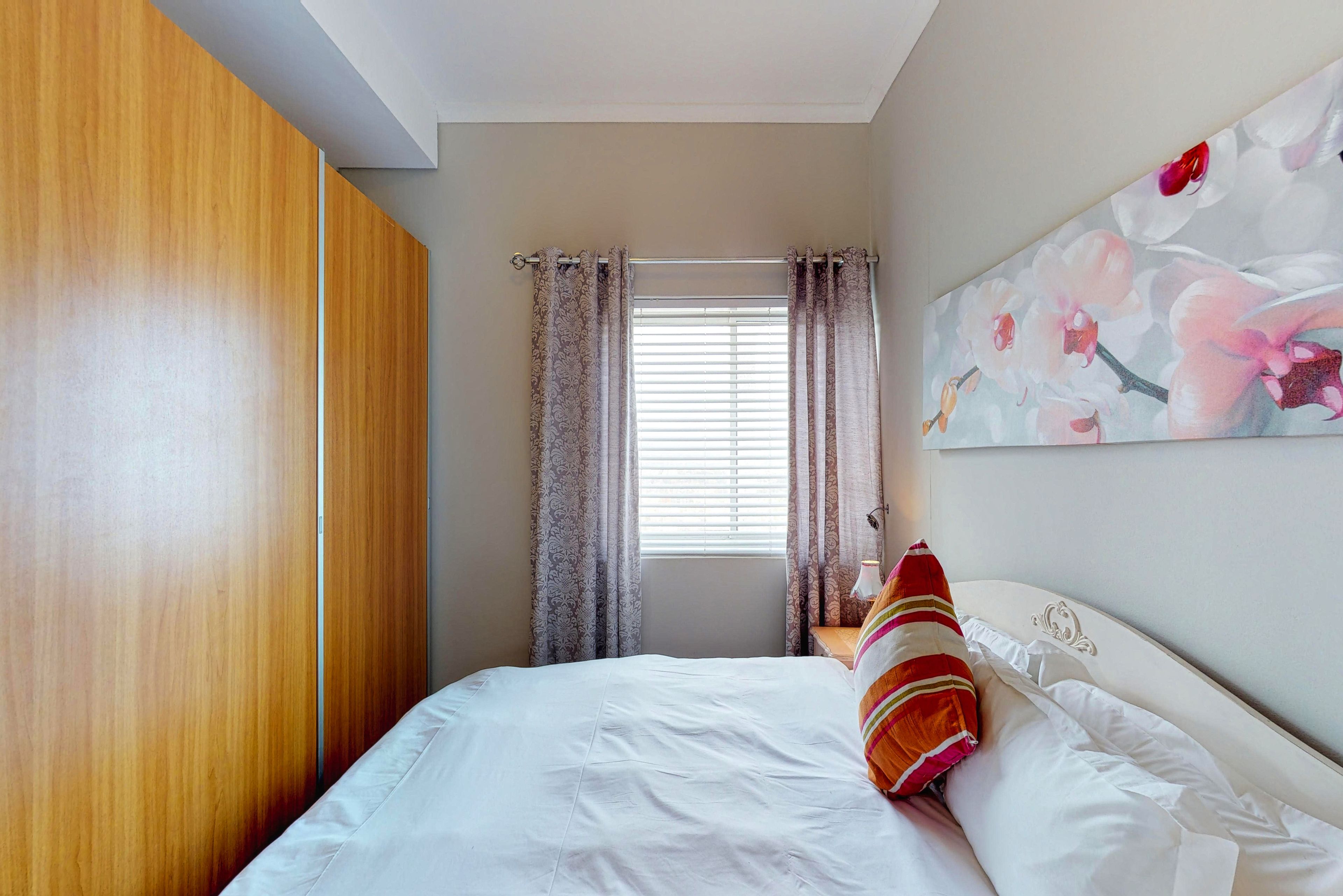 A bedroom with a bed, curtains, a window, and a wall with a floral painting.