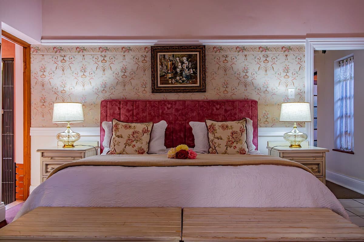 A bedroom with a large bed, floral-patterned wallpaper, a red upholstered headboard, two bedside tables with lamps, and a framed picture above the bed.