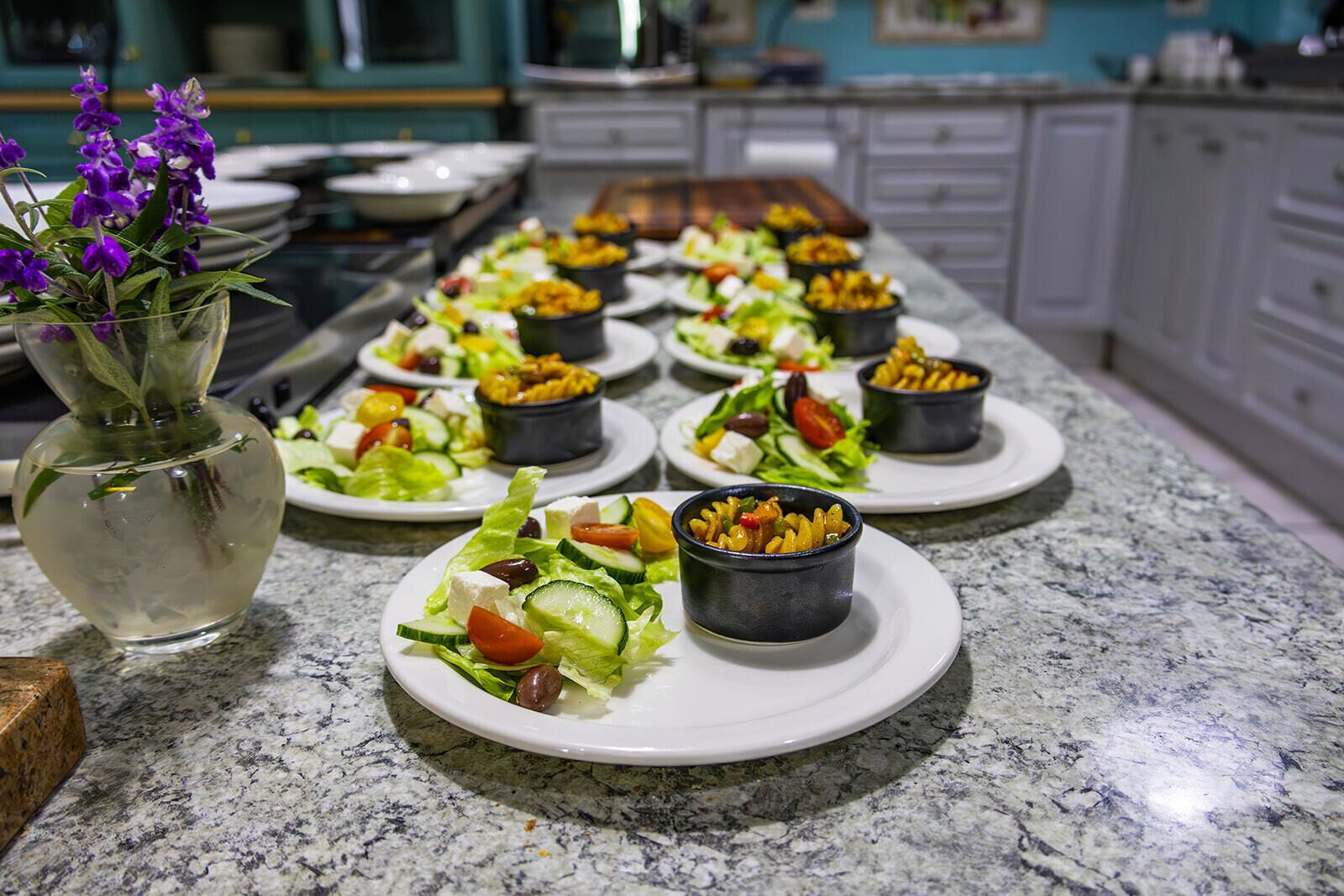 A kitchen counter with multiple plates of food, including salads and bowls of pasta, and a vase with purple flowers.