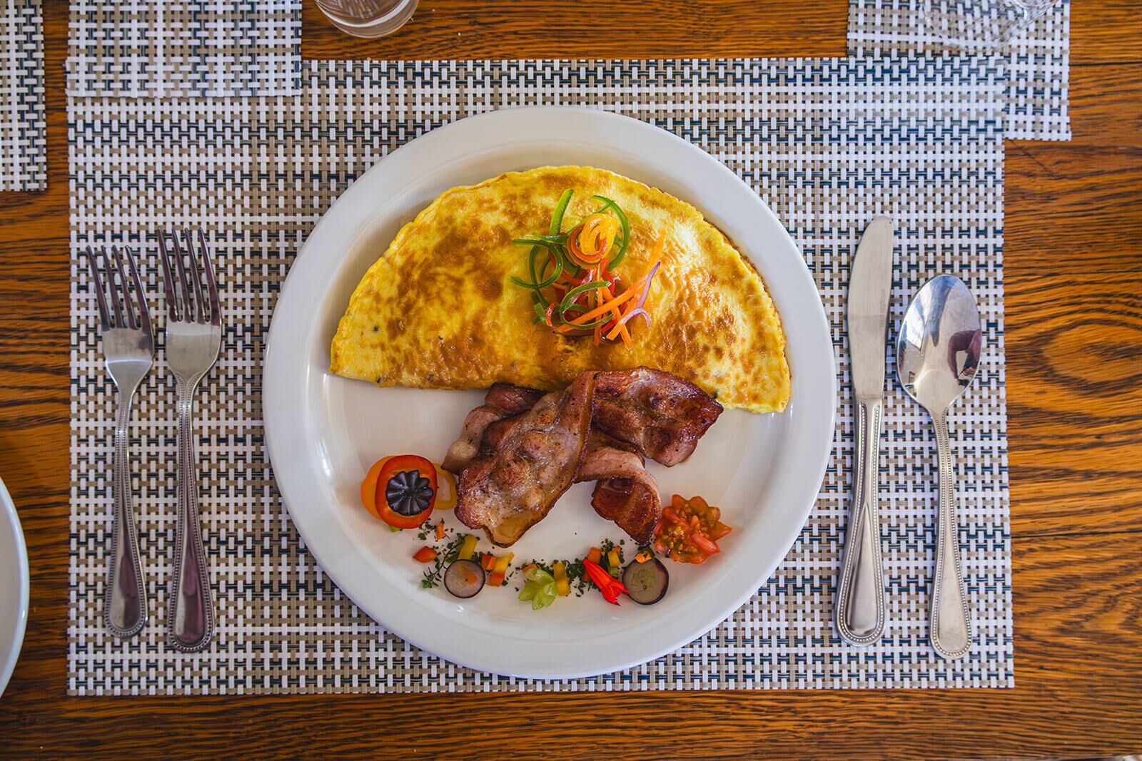 Un plato con una tortilla, tocino y verduras sobre un posavasos tejido.