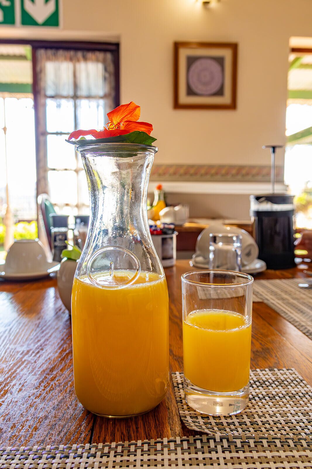 A glass pitcher of orange juice with a flower on top, placed on a wooden table with a glass beside it.