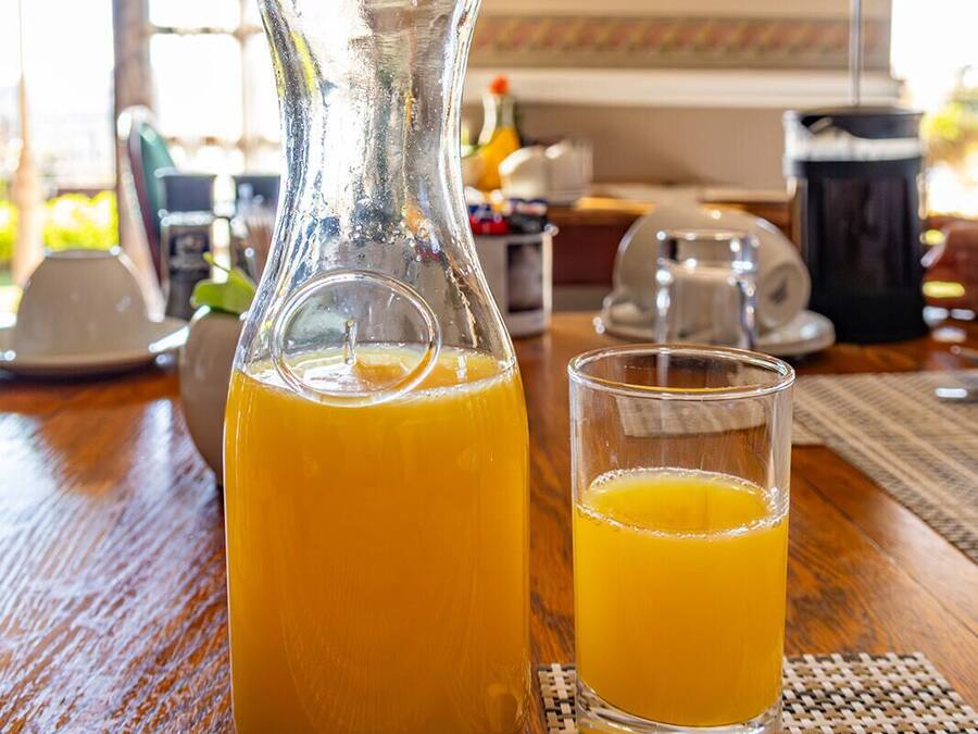 A glass pitcher of orange juice with a flower on top, placed on a wooden table with a glass beside it.