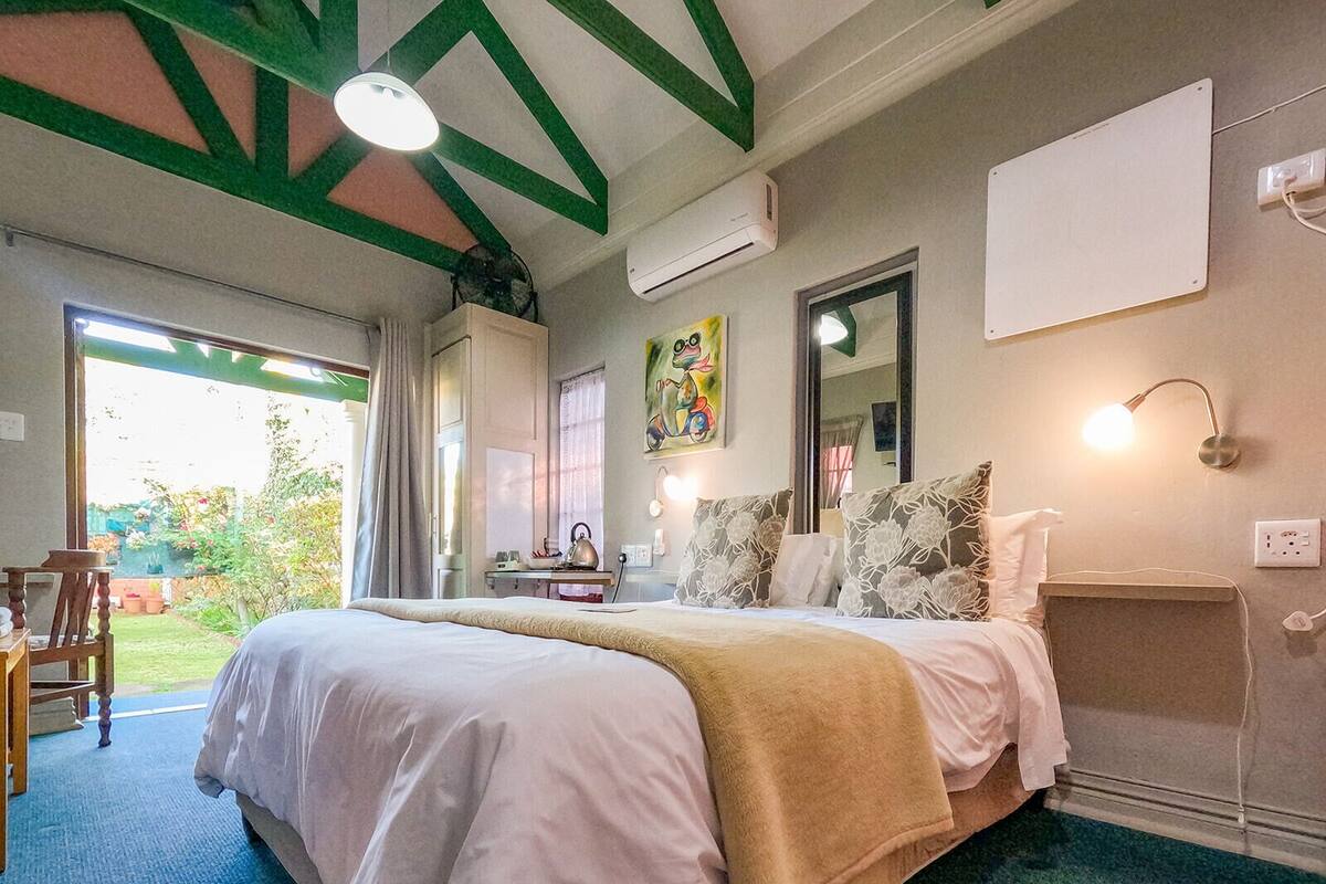 A bedroom with a large bed, a desk, and a chair. There is a window with curtains, a mirror, and a wall-mounted light.