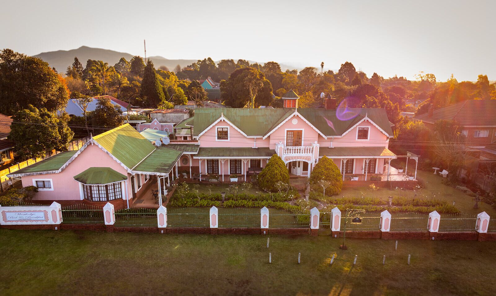 A large pink building with a green roof, surrounded by a well-maintained garden and a fence.